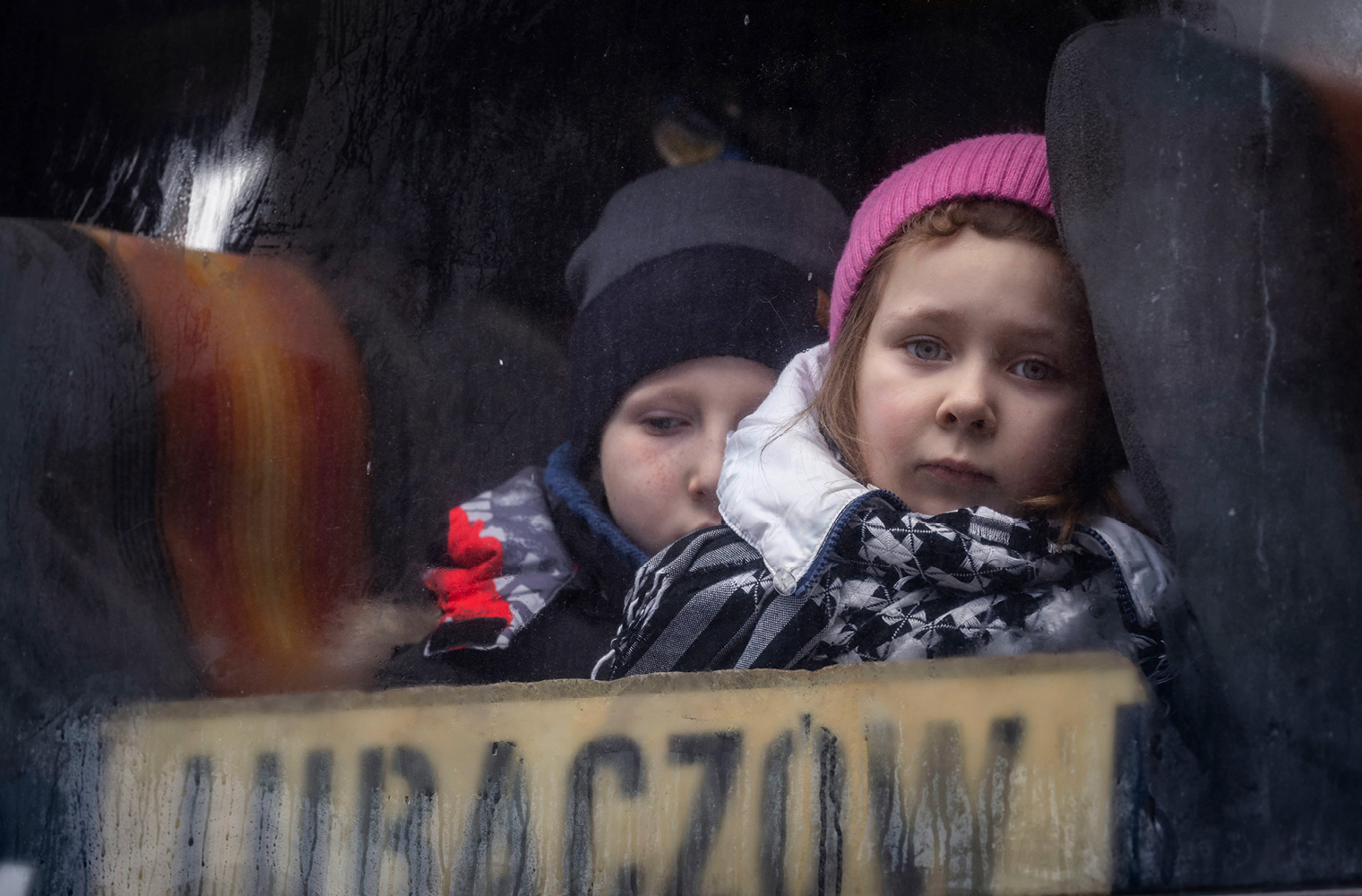 Das Bild zeigt zwei Kinder, die in einem Bus sitzen. Ihre Gesichtsausdrücke wirken traurig. Das Mädchen trägt eine pinke Haube und ist in eine dicke Jacke eingepackt. Der Bub trägt eine grau-schwarze Haube und trägt auch eine Jacke. 