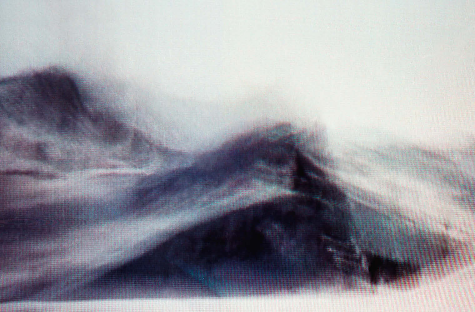 Ein Bild im Videostil zeigt eine Sand- oder Schneedüne, die über einen Gipfel fegt. Der Himmel ist weiß.
