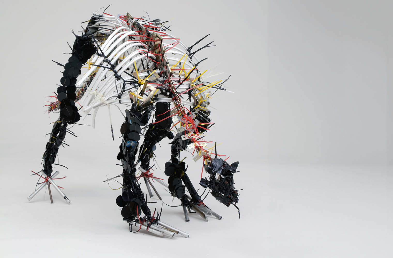 Skulptur eines Dinosauriers aus verschiedenen Materialien wie Kabel und Plastik in Schwarz, Weiß, Rot und Gelb. Die dynamische Haltung des Dinosauriers suggeriert Bewegung. Der neutrale Hintergrund betont die Details der komplexen Struktur.