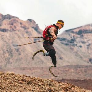 Ein Sportler mit Prothesen an beiden Beinen springt mit Wanderstöcken über steiniges Gelände. Er trägt eine rote Sportweste und eine Sonnenbrille. Im Hintergrund sind unscharfe Bergkonturen sichtbar.