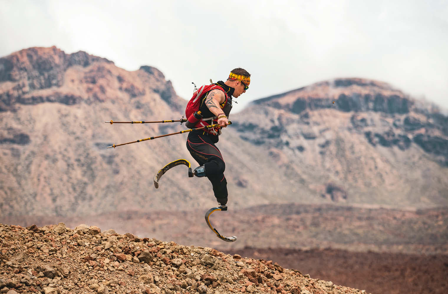 Ein Sportler mit Prothesen an beiden Beinen springt mit Wanderstöcken über steiniges Gelände. Er trägt eine rote Sportweste und eine Sonnenbrille. Im Hintergrund sind unscharfe Bergkonturen sichtbar.