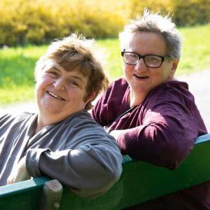 Zwei Frauen im mittleren Alter sitzen auf einer Parkbank und lächeln entspannt in die Kamera.