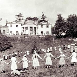 Auf einer leicht abfallenden Wiese vor dem Camphill-Haus stehen viele Frauen, Männer und Kinder in einem großen Kreis und halten sich an den Händen.