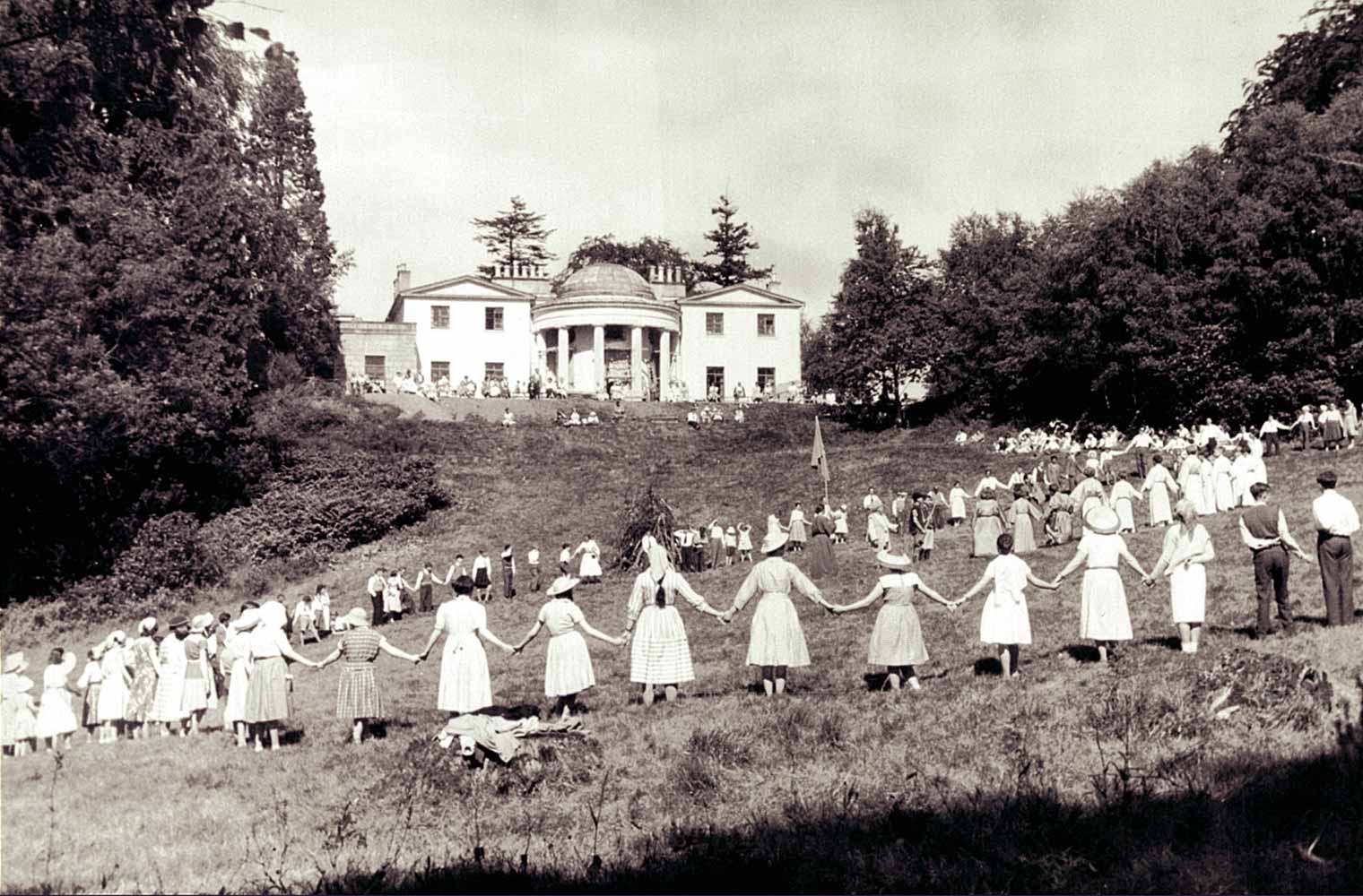 Auf einer leicht abfallenden Wiese vor dem Camphill-Haus stehen viele Frauen, Männer und Kinder in einem großen Kreis und halten sich an den Händen.