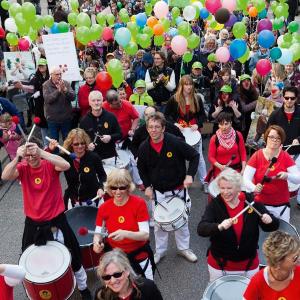 Auf einer Straße sind viele Menschen mit Trommeln, Luftballons und Transparenten unterwegs.