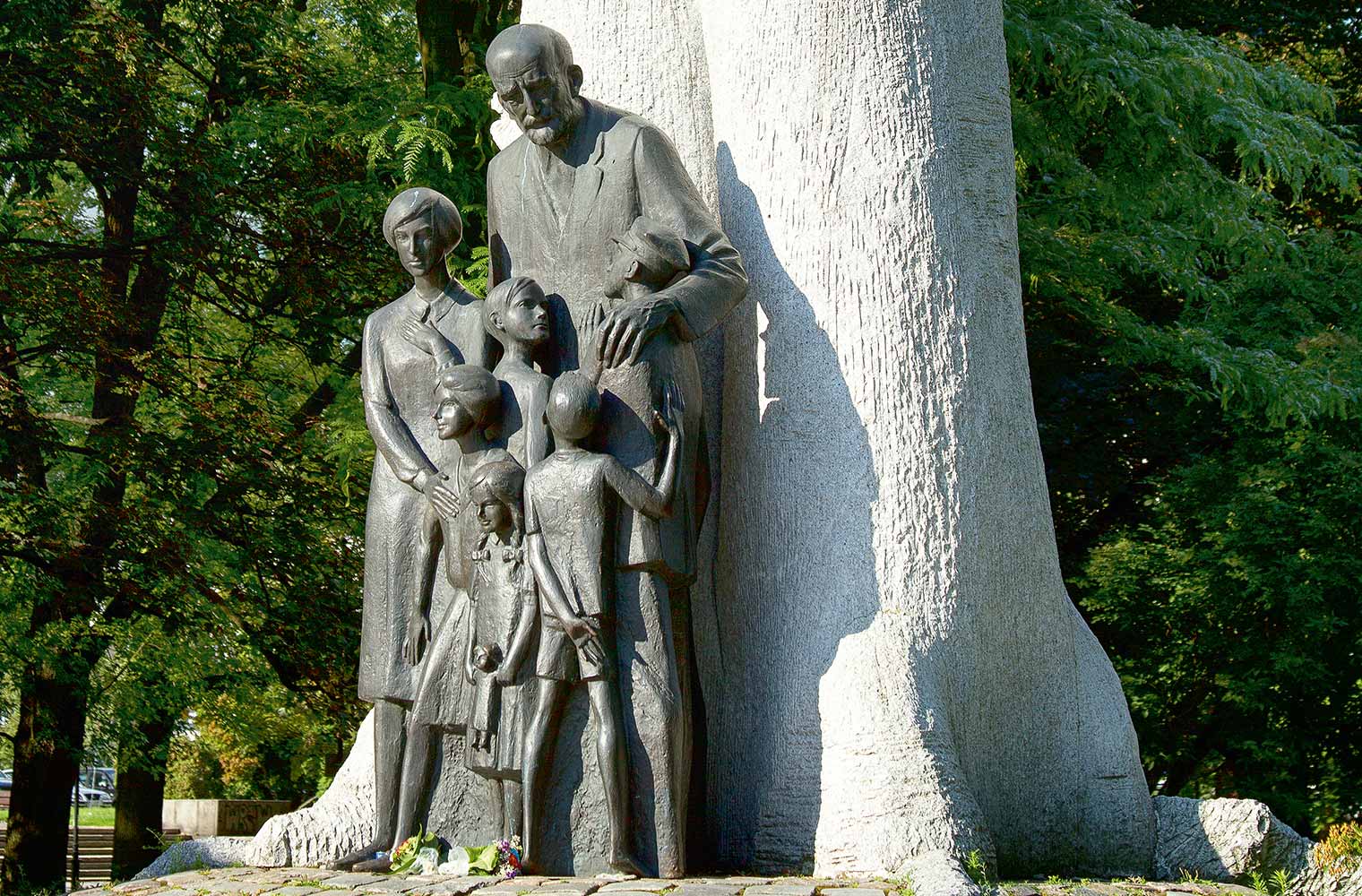  	Das Denkmal zeigt Janusz Korczak. Er steht vor einem großen Baum und hält seine Hände schützend um sechs Kinder unterschiedlichen Alters.