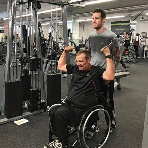 Der ehemalige Rollstuhlbasketballer Dirk Schmitz übt an einem Fitnessgerät. Sein Trainer Thomas Gessler steht dahinter.