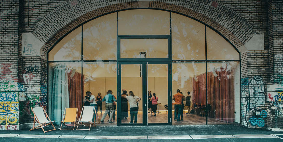 Die Aufnahme zeigt die Fassade eines Backsteingebäudes mit einem großen, bogenförmigen Fenster- und Türelement. Durch die Glasfront sind mehrere  P...