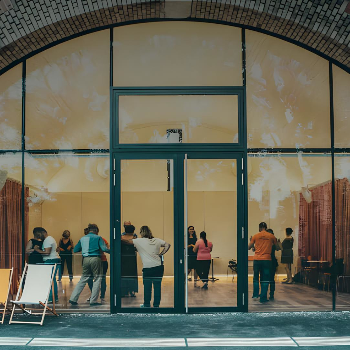 Die Aufnahme zeigt die Fassade eines Backsteingebäudes mit einem großen, bogenförmigen Fenster- und Türelement. Durch die Glasfront sind mehrere  P...