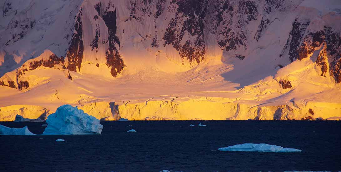 Ein großer Gletscher der Antarktis glänzt golden im Licht der Abendsonne. Dahinter befindet sich ein verschneiter Berg. Davor sieht man dunkelblaue...