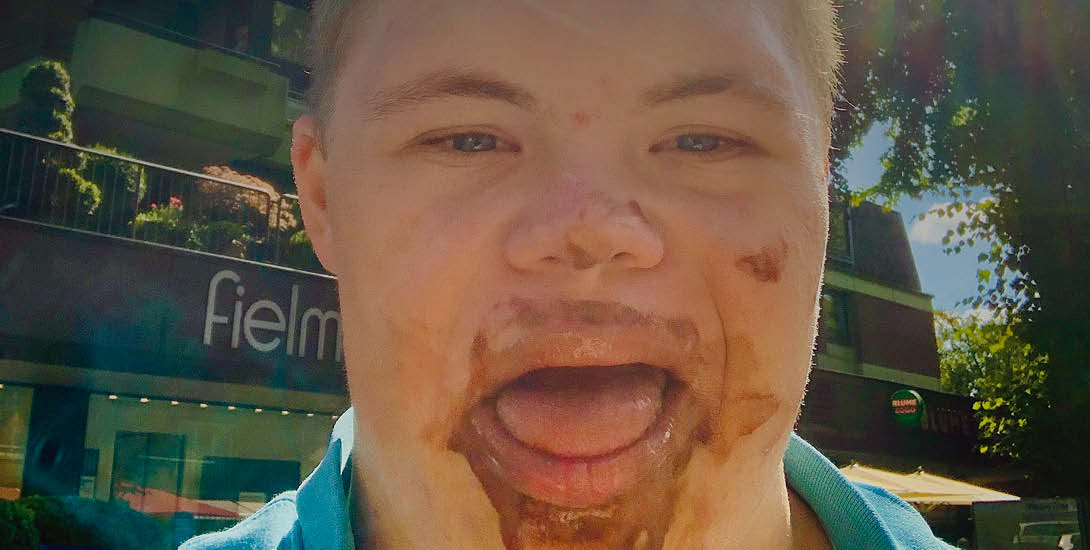 Ein junger Mann ist frontal im Freien fotografiert und blickt mit geöffnetem Mund in die Kamera. Im Mund- und Kinnbereich sind braune Verschmutzung...