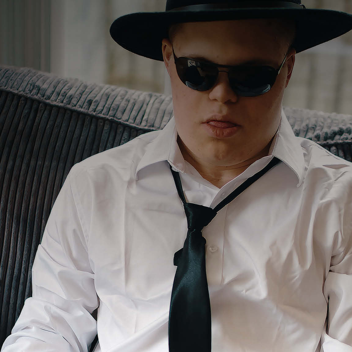 Ein junger Mann mit heller Haut sitzt auf einem Sofa. Er trägt ein weißes Hemd, eine schwarze Krawatte, eine schwarze Sonnenbrille und einen schwar...
