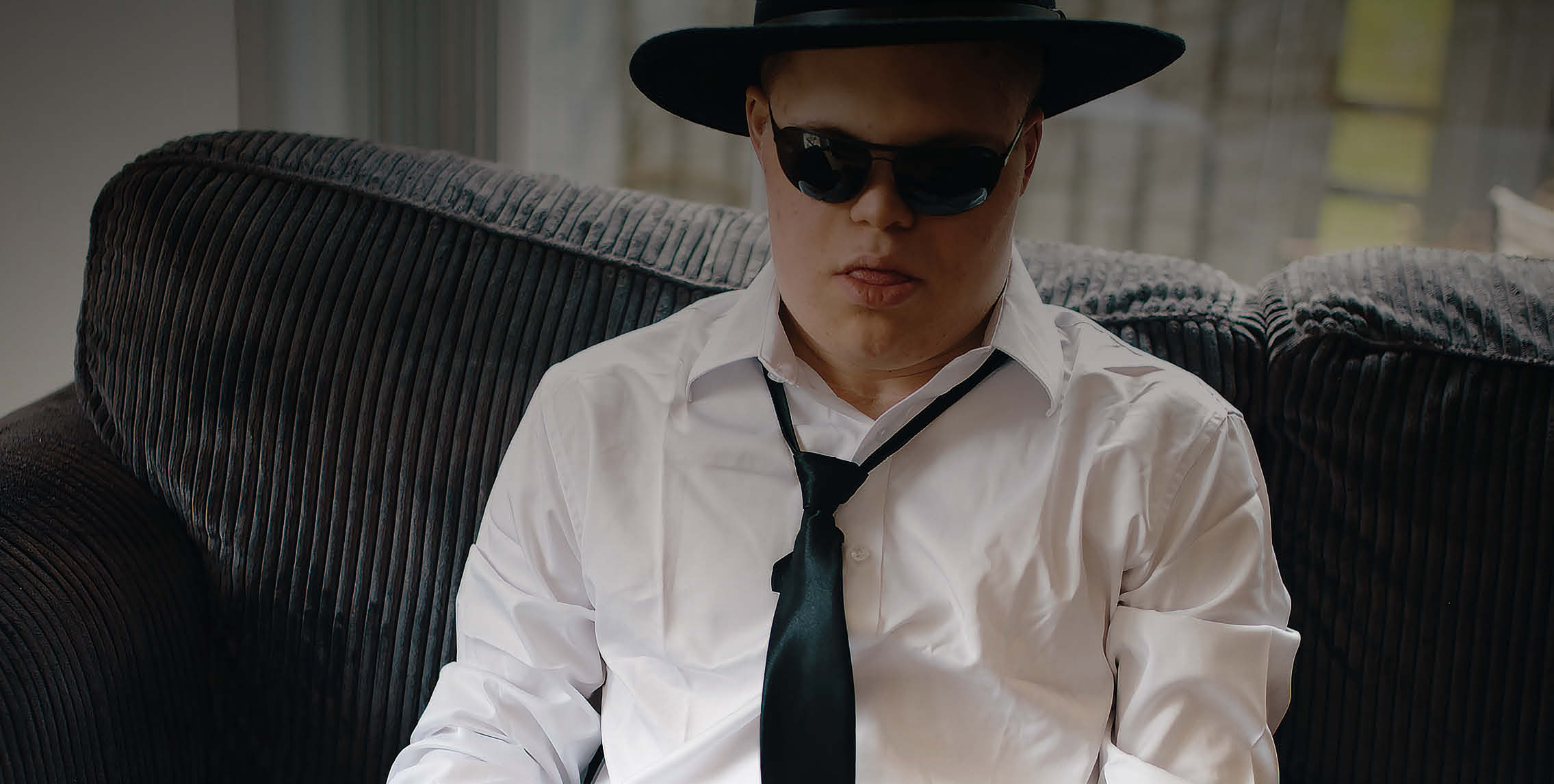 Ein junger Mann mit heller Haut sitzt auf einem Sofa. Er trägt ein weißes Hemd, eine schwarze Krawatte, eine schwarze Sonnenbrille und einen schwar...