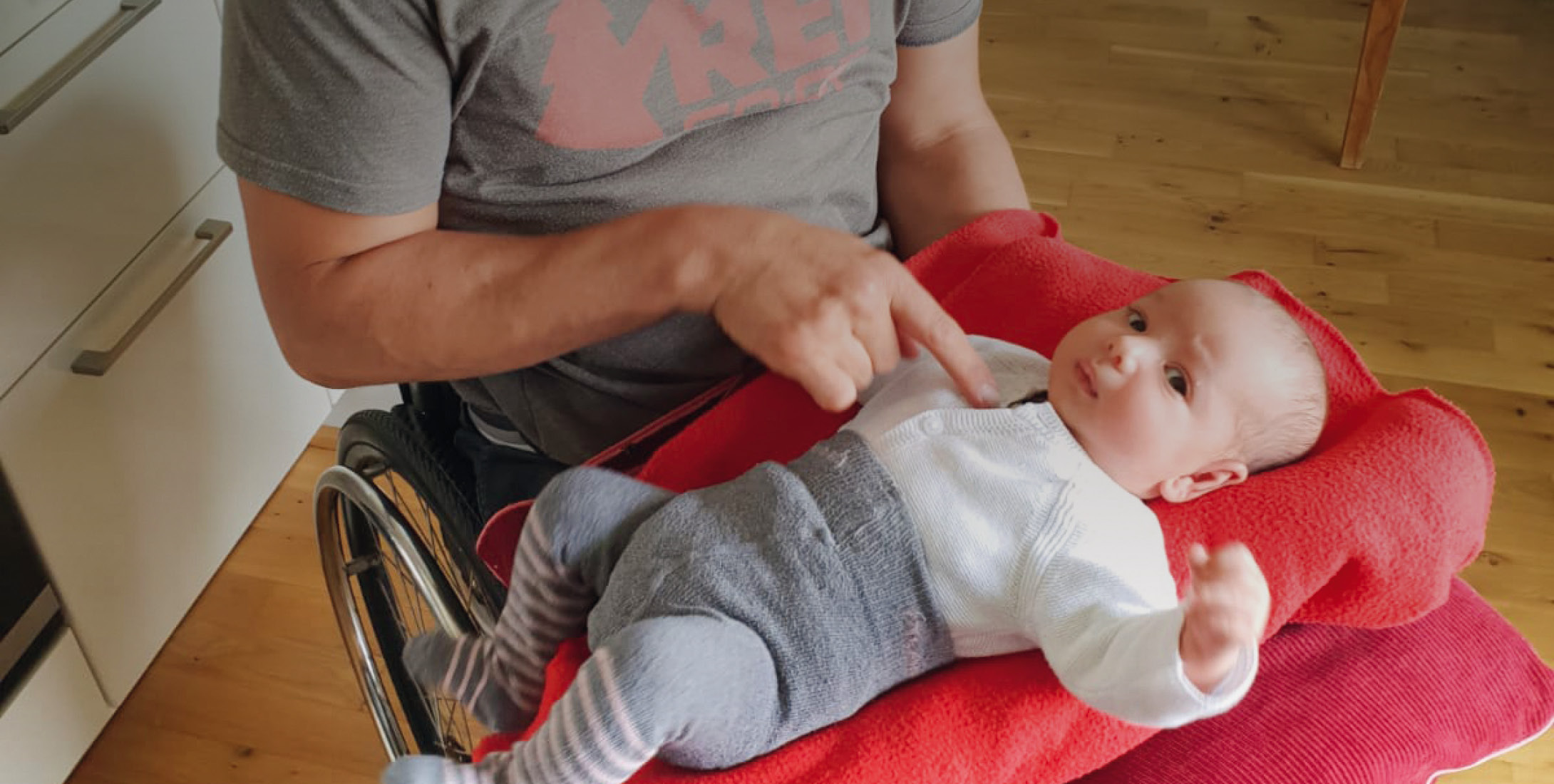 Ein lächelnder Mann sitzt im Rollstuhl in einem Wohnraum. Auf seinem Schoß befindet sich eine rote Decke, worauf ein Baby mit weißem Shirt und grau...