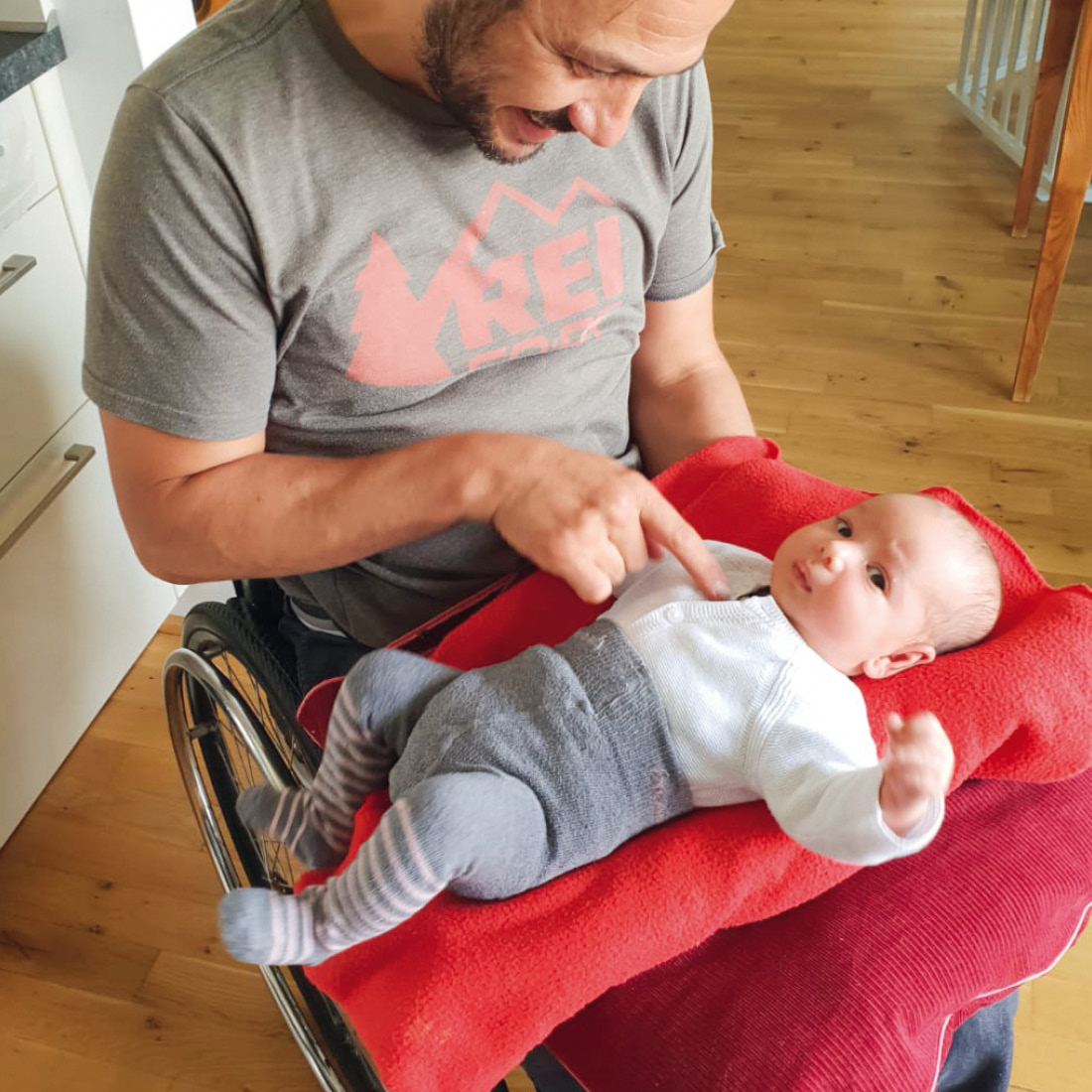 Ein lächelnder Mann sitzt im Rollstuhl in einem Wohnraum. Auf seinem Schoß befindet sich eine rote Decke, worauf ein Baby mit weißem Shirt und grau...