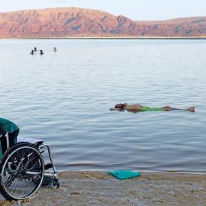 Ein leerer Rollstuhl steht am Strand. Im nahen Wasser genießt Reinfried Blaha rückenliegend das salzbedingte Schweben im Toten Meer.
