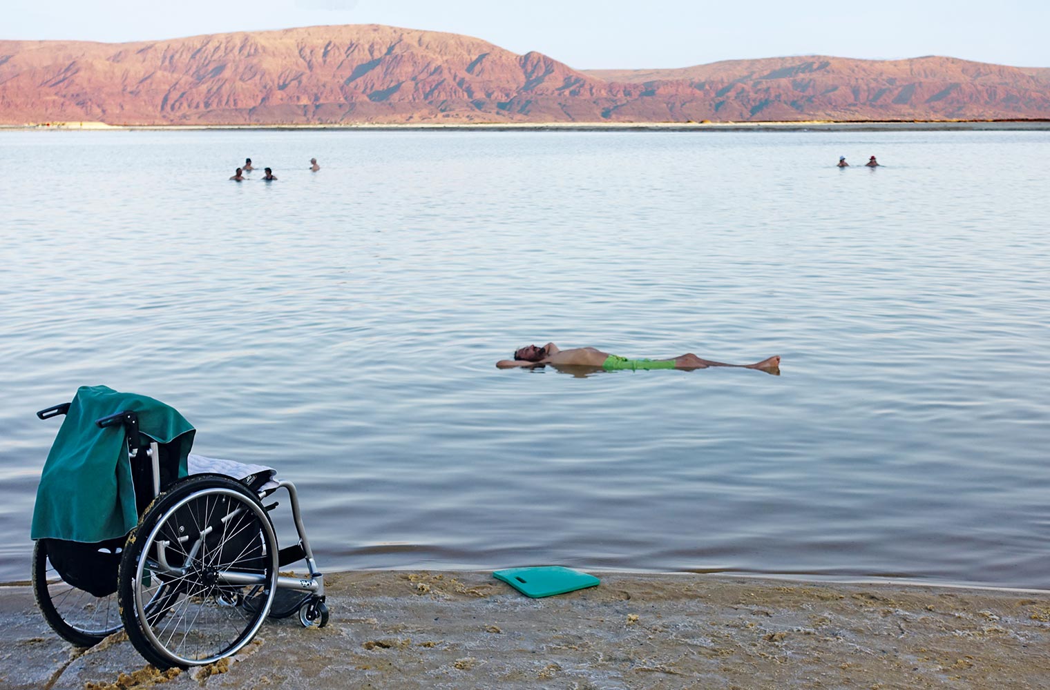 Ein leerer Rollstuhl steht am Strand. Im nahen Wasser genießt Reinfried Blaha rückenliegend das salzbedingte Schweben im Toten Meer.