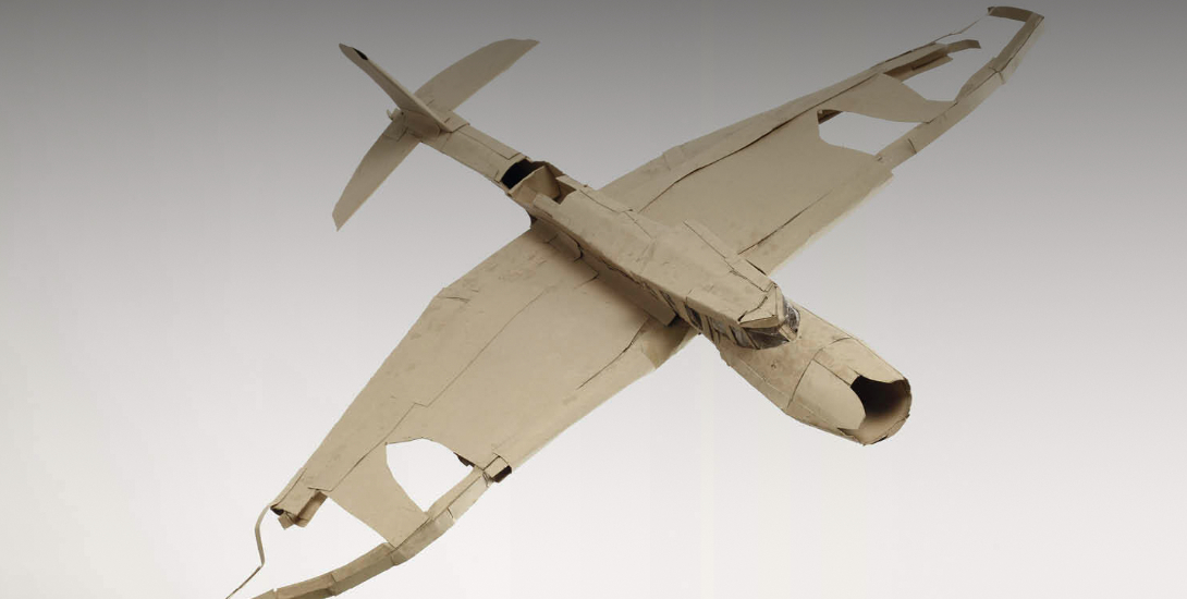 Ein Modellflugzeug aus Pappe mit ausgebreiteten Flügeln. Die Oberfläche wirkt uneben, die Kanten sind geknickt und geklebt. 