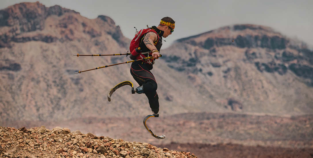 Ein Sportler mit Prothesen an beiden Beinen springt mit Wanderstöcken über steiniges Gelände. Er trägt eine rote Sportweste und eine Sonnenbrille....