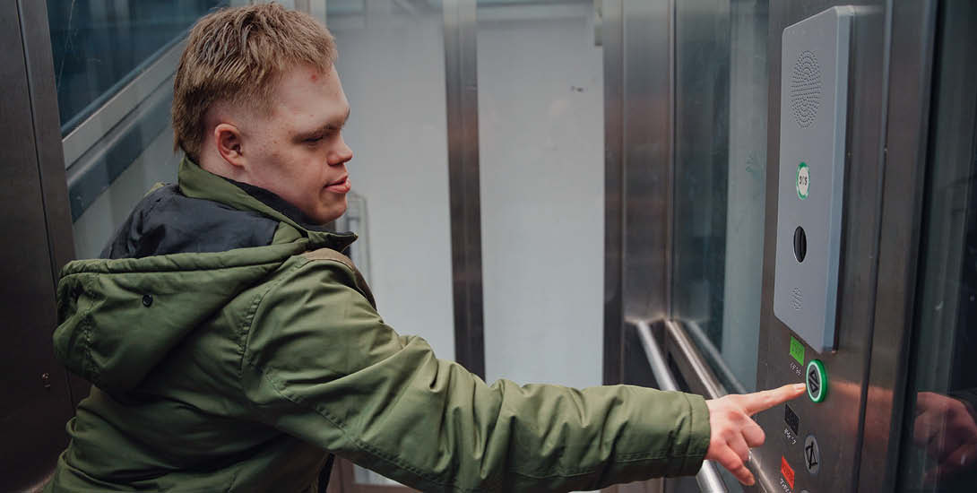 Eine Person in einer grünen Jacke steht in einem Aufzug und drückt mit ausgestrecktem Arm einen Knopf an der Bedienleiste. Die Aufzugskabine hat Me...