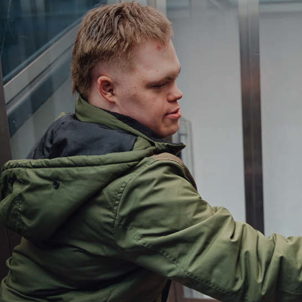 Eine Person in einer grünen Jacke steht in einem Aufzug und drückt mit ausgestrecktem Arm einen Knopf an der Bedienleiste. Die Aufzugskabine hat Me...