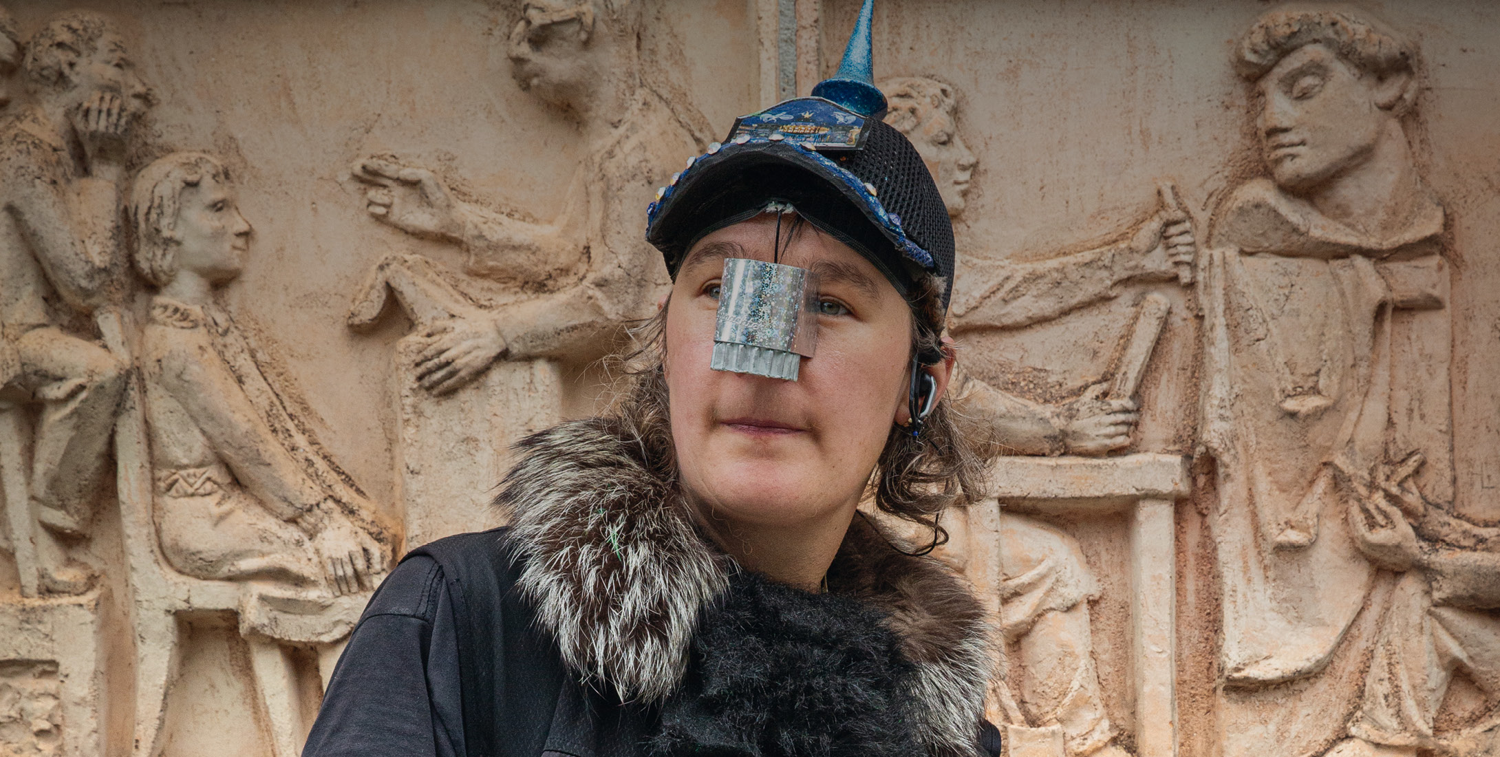 Eine Person trägt eine schwarze Kopfbedeckung mit blauem, spitzem Aufsatz. Auf der Nase befindet sich ein silberfarbenes, rechteckiges Element. Im...