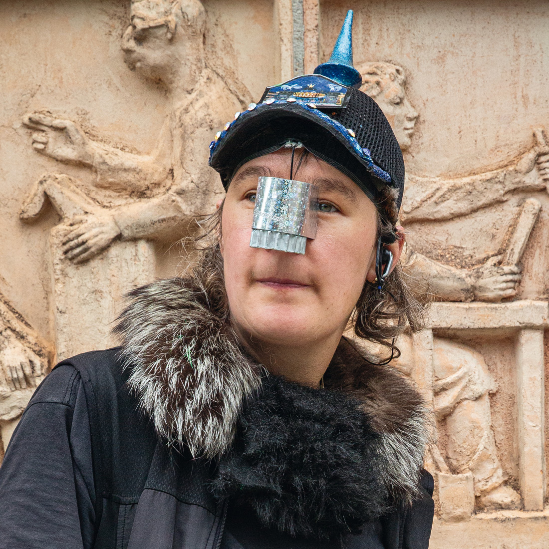 Eine Person trägt eine schwarze Kopfbedeckung mit blauem, spitzem Aufsatz. Auf der Nase befindet sich ein silberfarbenes, rechteckiges Element. Im...