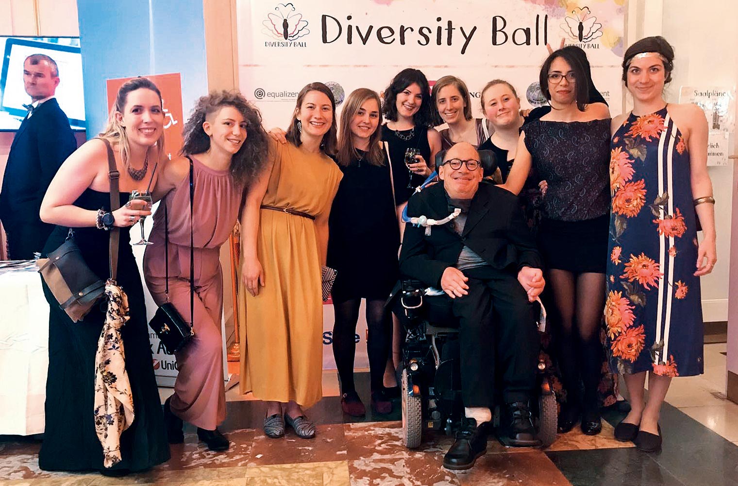 Franz-Joseph Huainigg sitzt festlich gekleidet lächelnd in seinem Rollstuhl umgeben von Assistentinnen. Dahinter steht ein Plakat zum Diversity Ball.