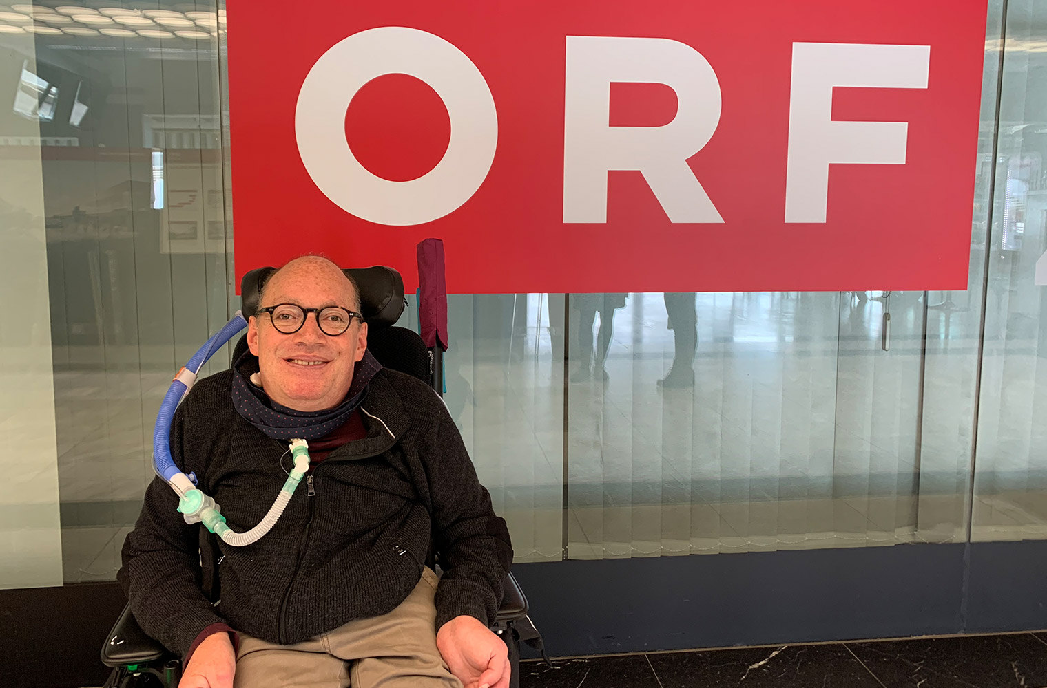 Franz-Joseph Huainigg sitzt lächelnd in seinem Rollstuhl vor dem ORF-Gebäude, in dem er arbeitet. Sein Beatmungsschlauch ist unaufdringlich erkennbar.