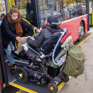 Ein Mann in einem E-Rolli rollt gerade über eine Rampe in einen Bus der Wiener Linien. Seine Persönliche Assistenzin hilft ihm dabei. 