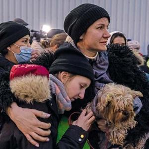 Eine Frau trägt ihren Hund mit der einen Hand und mit dem anderen Arm hält sie ein Mädchen. Beide tragen eine Jacke, Haube und einen Schal. Das Mädchen trägt auch noch einen pink karierten Rucksack. Im Hintergrund sind weitere Massen an Menschen zu sehen. 