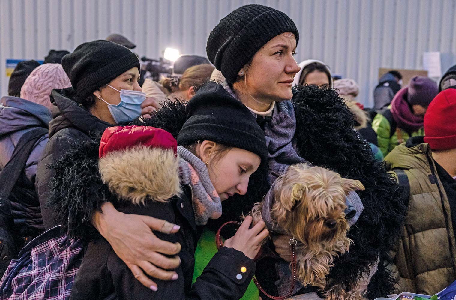 Eine Frau trägt ihren Hund mit der einen Hand und mit dem anderen Arm hält sie ein Mädchen. Beide tragen eine Jacke, Haube und einen Schal. Das Mädchen trägt auch noch einen pink karierten Rucksack. Im Hintergrund sind weitere Massen an Menschen zu sehen. 