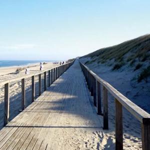 Ein zum Teil sandbedeckter Steg ist zu erkennen. Links davon ein aufgeschütteter Hang mit vereinzelten Grasbüscheln. Rechts davon ein Sandstrand und anschließend das blaue Meer. 