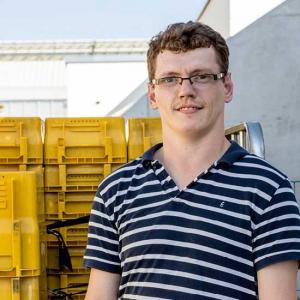 Ein junger hellhäutiger Mann mit blonden Haaren, Brille und einem weiß-blau gestreiftem T-Shirt steht vor einem Stapel gelber Postcontainer im Freien. Im Hintergrund sieht man ein weißes Gebäude und einen blauen Himmel. 