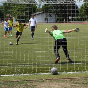 Ein Fußballspiel ist im Gange. Mehrere Spieler sind am Feld und ein Stürmer will gerade auf das Tor, in welchem ein Jugendlicher mit grünem Trikot steht, schießen. Der Rasen ist saftig grün und im Hintergrund sieht man grüne Bäume. Der Himmel ist blau. 