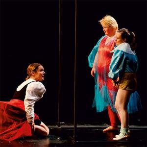 Eine Frau mittleren Alters kniet in ihrem weiß-schwarzen Kleid mit rotem Rock vor zwei jungen Frauen, welche moderne Kleider in rot-blau tragen. Eine Frau mittleren Alters kniet in ihrem weiß-schwarzen Kleid mit rotem Rock vor zwei jungen Frauen, welche moderne Kleider in rot-blau tragen. Der Hintergrund der Bühne ist schwarz.