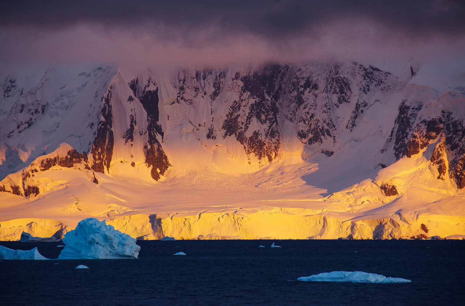 Ein großer Gletscher der Antarktis ist abgebildet. Im Vordergrund sieht man das dunkelblaue Meer und einen treibenden Eisberg. Auch etwas Schelfeis befindet sich im Wasser.  Der Himmel ist von einem hellgrauen Nebel bedeckt. 