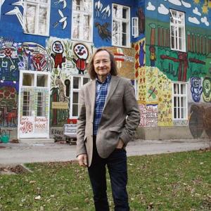 Ein Mann mittleren Alters mit langen braunen Haaren und Brille steht vor einem mit Graffiti bemalten Haus. Dieses ist mit vielen Motiven bemalt worden. Menschen, Schmetterlinge und abstrakte Formen können der Mauer entnommen werden. Vor dem Gebäude befindet sich ein asphaltierter Weg und eine Wiese. 