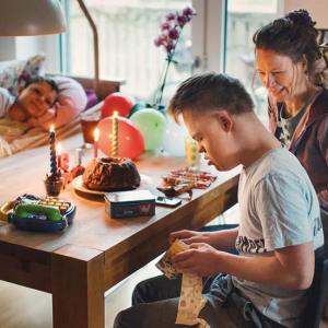 Eine Familie ist am Foto abgebildet. Eine Frau mittleren Alters mit brünetten Haaren sitzt am Esstisch und schaut zu ihrem Sohn, der gerade eine Packung öffnet. Der Junge hat dunkelblondes Haar und ein weißes kurzärmliges T-Shirt an. Er hat Trisomie-21. Auf dem schlichten, robusten Holztisch steht unter anderen Leckereien, ein Geburtstagsguglhupf mit ein paar angezündeten Kerzen. Gegenüber liegt ein Mädchen im Bett. Sie ist zugedeckt und schaut lächelnd zu ihrem Bruder. 