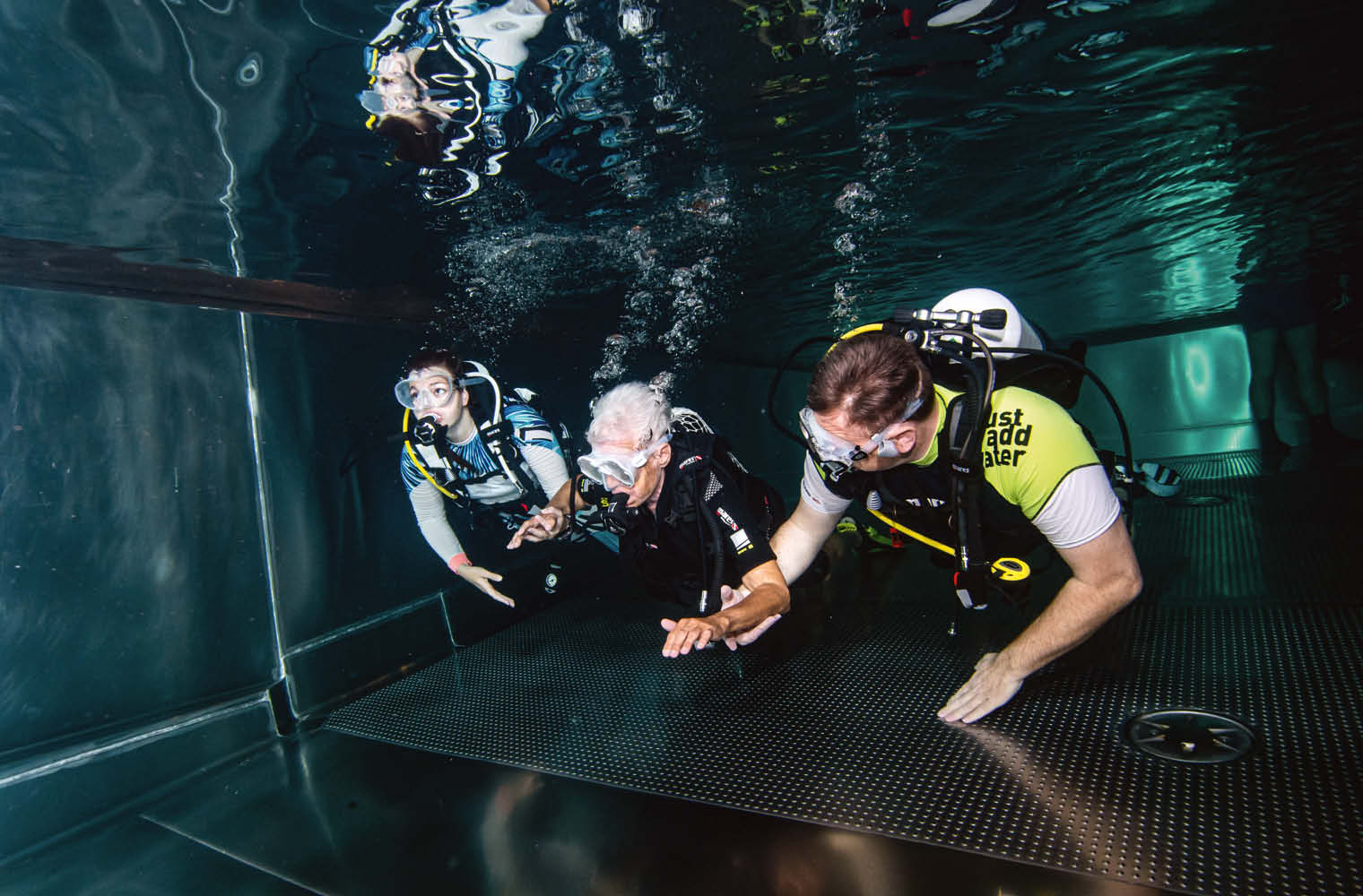 Ein Unterwasserfoto zeigt eine ältere Person und zwei jüngere Männer mit Tauchanzug und -brille, sowie Pressluftflasche unter Wasser am Grund eines Tauchbeckens. Es steigen Blubberblasen der Tauchenden auf. Das Wasser ist klar.