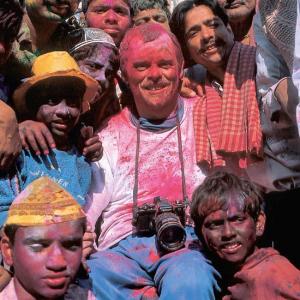 Ein lächelnder, im Rollstuhl sitzender Mann mit umgehängter Kamera, umgeben von einer Gruppe bunt beschmierter Menschen beim indischen Holi-Fest, strahlt Freude und Gemeinschaft aus. Farbpulver in Rosa und Blau bedeckt ihre Gesichter und Kleidung, was eine ausgelassene Stimmung vermittelt.
