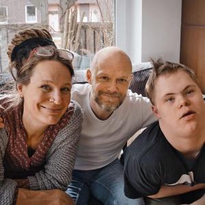 Ein fröhliches Familienporträt im Wohnzimmer, in dem eine Frau und ein Mann neben einem jungen Erwachsenen sitzen. Die lächelnde Frau trägt ein graues Strickoberteil. Der Mann, bärtig und kahlköpfig im weißen Shirt, lächelt ebenfalls und umarmt seine Frau und seinen erwachsenen Sohn mit Trisomie 21.