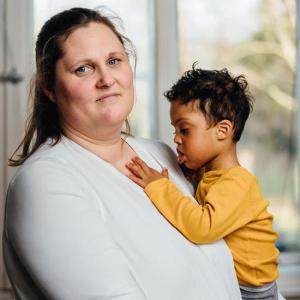 Eine Frau in einem weißen T-Shirt hält liebevoll ein kleines Kind, das ein gelbes Shirt trägt. Die Mutter blickt in die Kamera, während das Kind die Hand auf der Brust der Frau ruhen lässt. Beide stehen vor einem hellen Fenster, das sanftes Tageslicht in den Raum lässt und eine warme, ruhige Atmosphäre schafft.