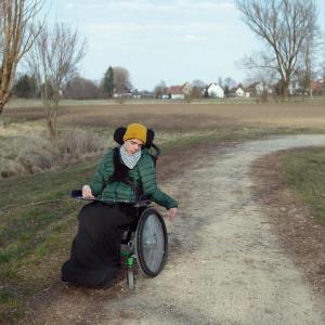 Eine Person in einem Rollstuhl, bekleidet mit einer grünen Jacke und einer gelben Mütze, befindet sich auf einem sandigen Weg in einer ländlichen Umgebung. Die Person blickt nachdenklich zur Seite, während die linke Hand neben dem Rollstuhl hinunterhängt. Im Hintergrund sind offene Felder und einige Häuser unter einem klaren Himmel zu sehen.