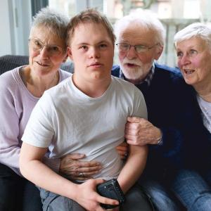 Ein Familienporträt zeigt einen jungen Mann mit Trisomie 21, umgeben von seinen Großeltern. Die Großmutter sitzt links neben ihm und lächelt ihn liebevoll an, während der Großvater und die zweite Großmutter ihn von der rechten Seite umarmen und ebenfalls lächeln. Die Szene spielt sich in einem hellen, wohnlichen Raum ab.
