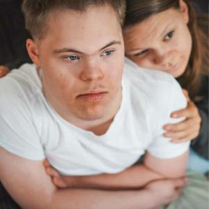 Ein junger Mann mit Trisomie 21 sitzt nachdenklich da, während eine Jugendliche ihn tröstend von der Seite umarmt. Der Junge trägt ein weißes T-Shirt und das Mädchen einen grauen Pullover und eine helle Hose.