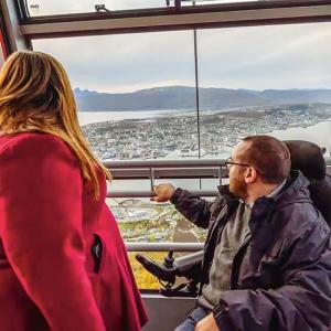 Eine Frau in einem roten Mantel und ein Mann im Rollstuhl mit schwarzer Jacke blicken gemeinsam aus einer verglasten Gondel auf eine weitläufige Stadtlandschaft mit Bergen und Wasser im Hintergrund. Der Mann deutet mit seiner Hand nach draußen, während beide die Aussicht genießen.