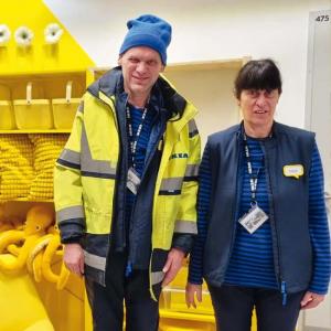 Zwei Mitarbeitende von IKEA stehen vor einer gelben Wand mit Regalen, die mit gelben Stofftieren und Kissen gefüllt sind. Beide tragen Arbeitskleidung mit Namensschildern, der Mann zusätzlich eine gelbe Jacke und eine blaue Mütze. Im Hintergrund sind gelbe Möbel und Dekorationen zu sehen, die das Bild farblich dominieren.