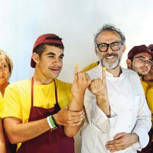 Eine Gruppe von Menschen in gelben T-Shirts und roten Schürzen steht nebeneinander. In der Mitte zeigt ein Mann im weißen Kochmantel mit einem jungen Mann Nudelteigstücke auf den Fingern. Alle lachen und wirken fröhlich.