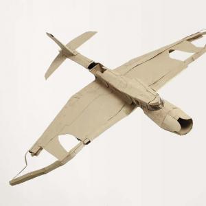 Ein Modellflugzeug aus Pappe mit ausgebreiteten Flügeln. Die Oberfläche wirkt uneben, die Kanten sind geknickt und geklebt. Das Flugzeug ist vor hellem Hintergrund fotografiert.